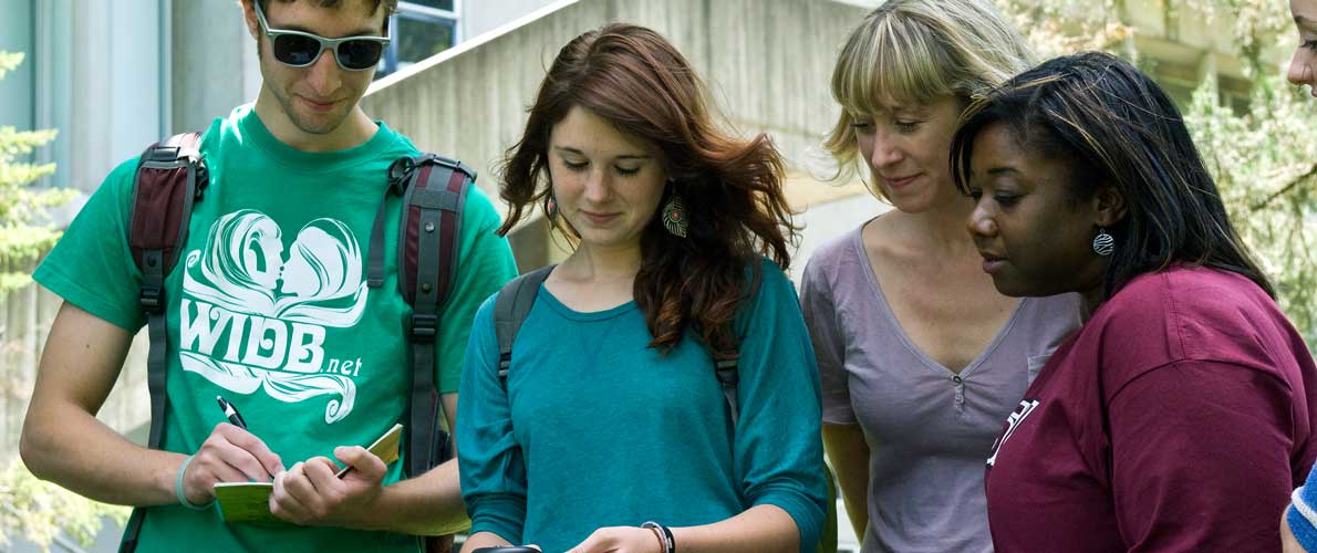 SIU Students standing together outdoors on campus, smiling and looking at a digital tablet held by the student in the center.